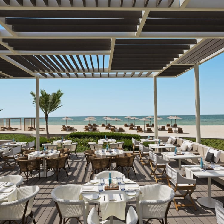 Outdoor-Restaurantbereich mit Tischen unter einem beschatteten Pergola, mit Blick auf einen Strand mit Liegestühlen und Sonnenschirm, im Hintergrund Palmen.