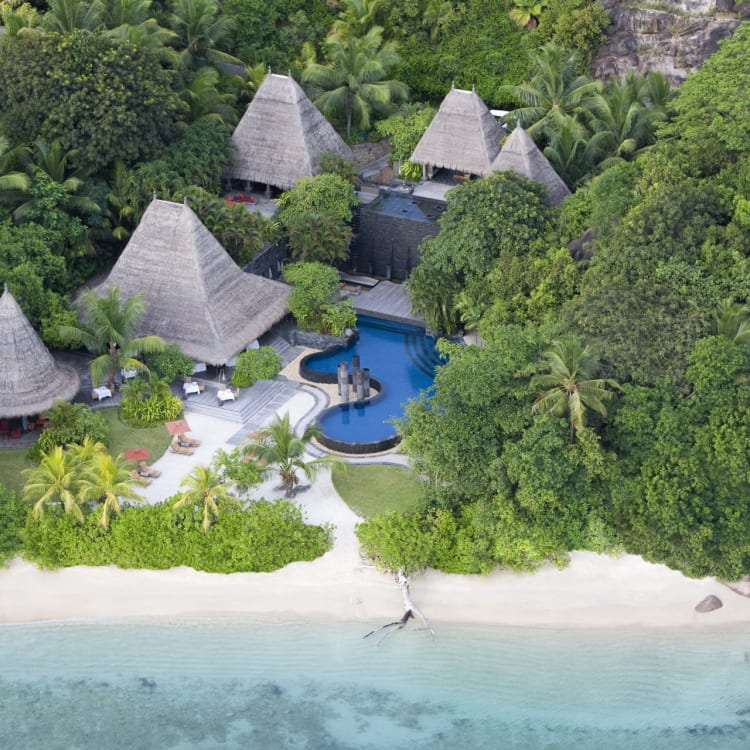 Luftaufnahme eines tropischen Resorts mit Thataghütten, einem Swimmingpool und einem weißen Sandstrand, umgeben von üppigem Grün.