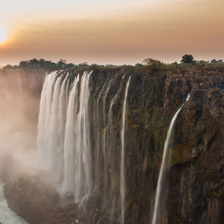Panoramablick auf die Viktoriafälle bei Sonnenuntergang mit Wasserfällen und aufsteigendem Nebel vom Fluss.