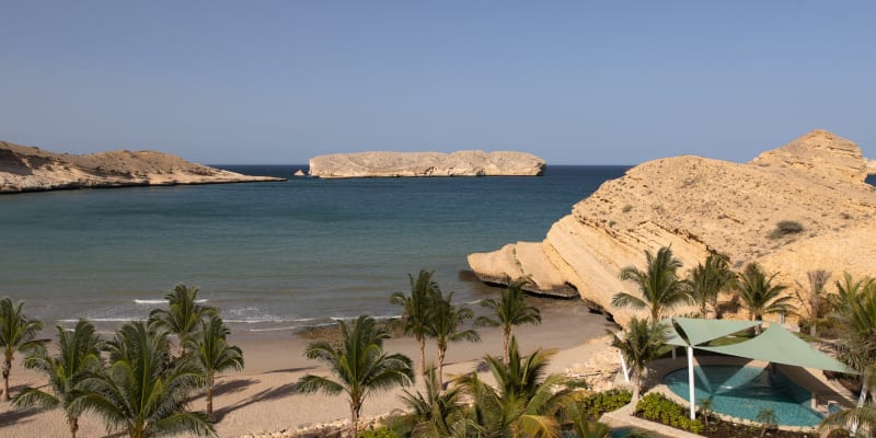 Luxury resort with private pools and palm trees overlooking a sandy beach and rocky islands in the ocean.