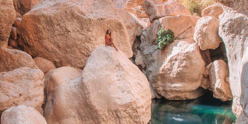 Eine Person sitzt auf einem großen Felsen in einer engen Schlucht mit ruhigem, spiegelndem Wasser und felsigen, rötlichen Felsen.