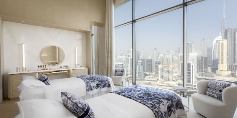 Elegant hotel room with two neatly made beds, large floor-to-ceiling windows overlooking a city skyline, and modern decor.