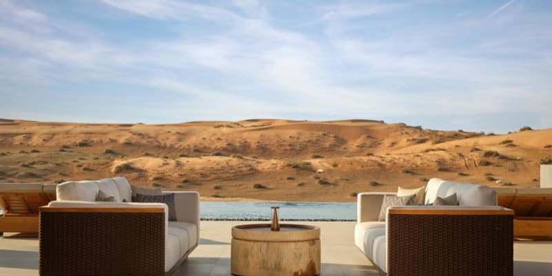Two sofas facing each other with a small round table between them on a terrace overlooking desert dunes and a river under a partly cloudy sky.