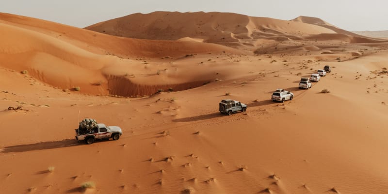 Eine Fahrzeugkolonne fährt durch eine weite Wüste mit Sanddünen in Oman.