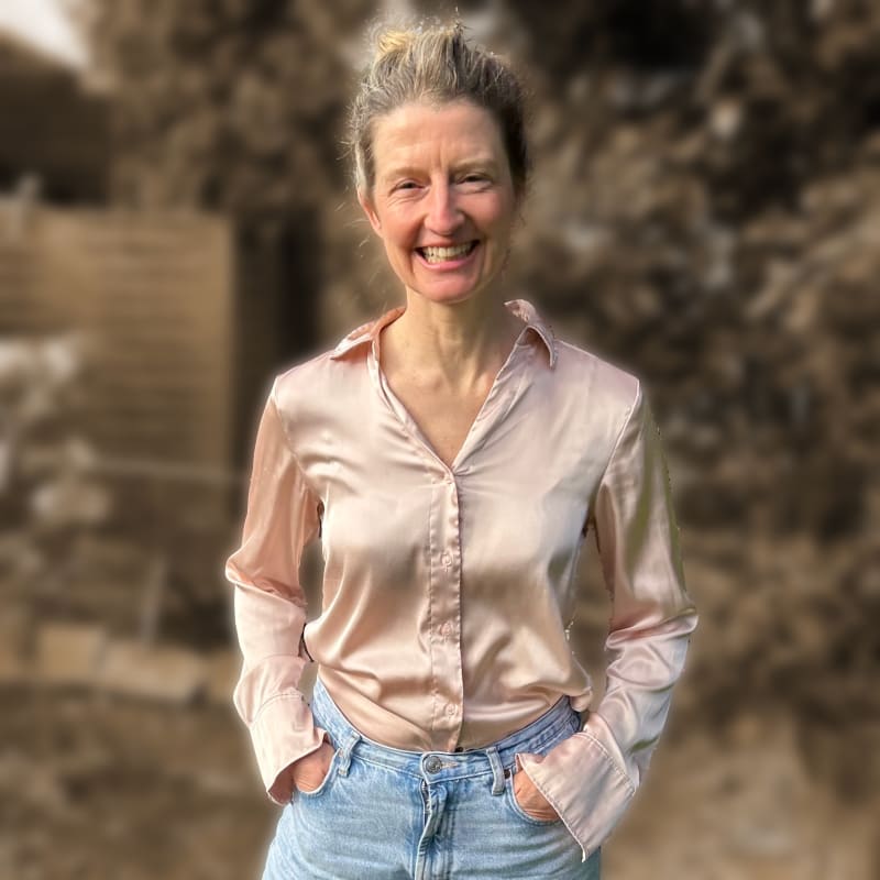 A smiling woman with gray hair tied back, wearing a silky beige blouse and blue jeans, standing outdoors with a blurred natural background.