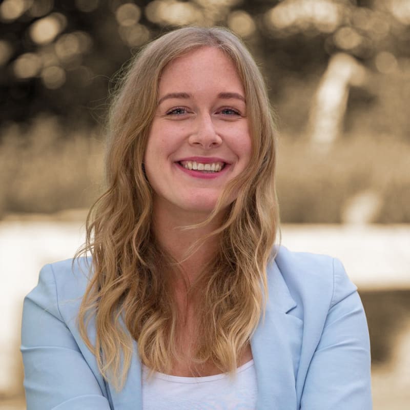 A young woman with long wavy blonde hair smiling outdoors, wearing a light blue blazer.