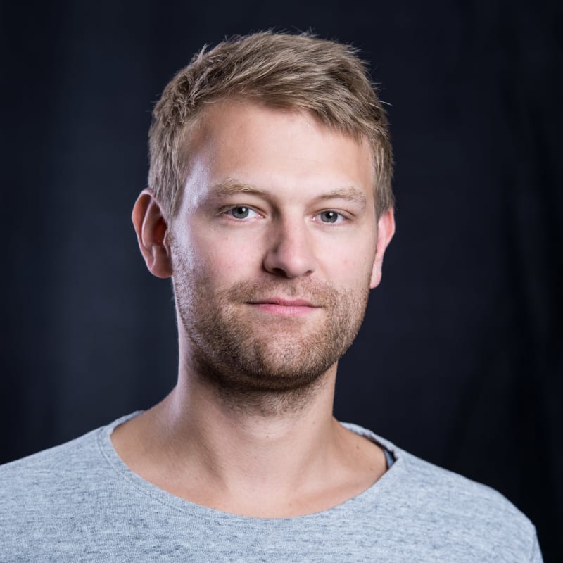 A young man with light brown hair and facial hair, wearing a light gray shirt, posed against a dark background.