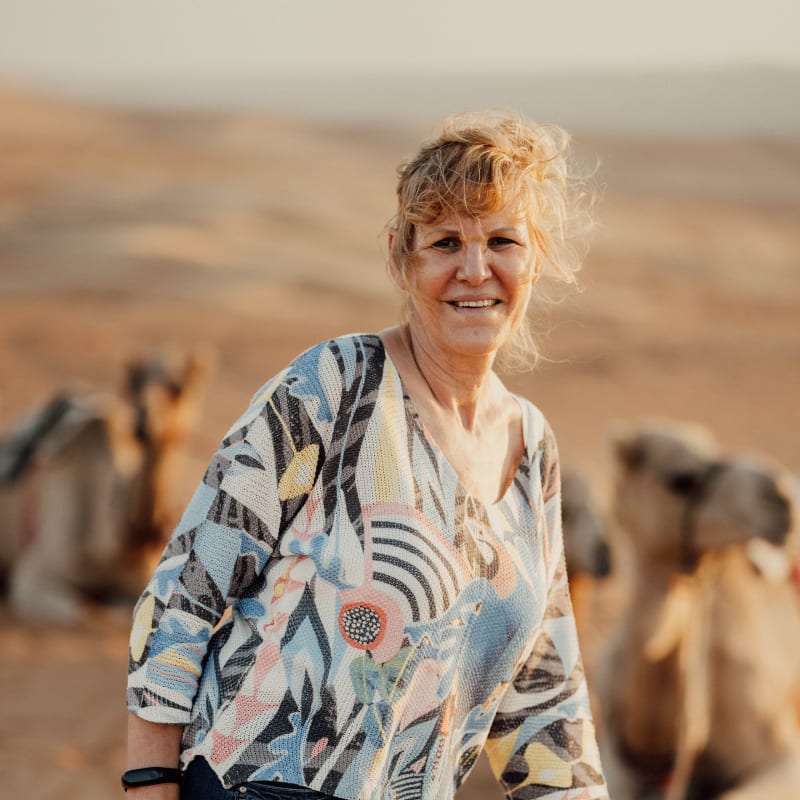 A smiling woman standing in a desert landscape with camels in the background during sunset.