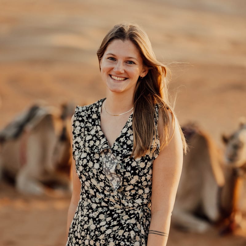 A young woman with long brown hair smiling outdoors in a sunny, rural setting with camels in the background.