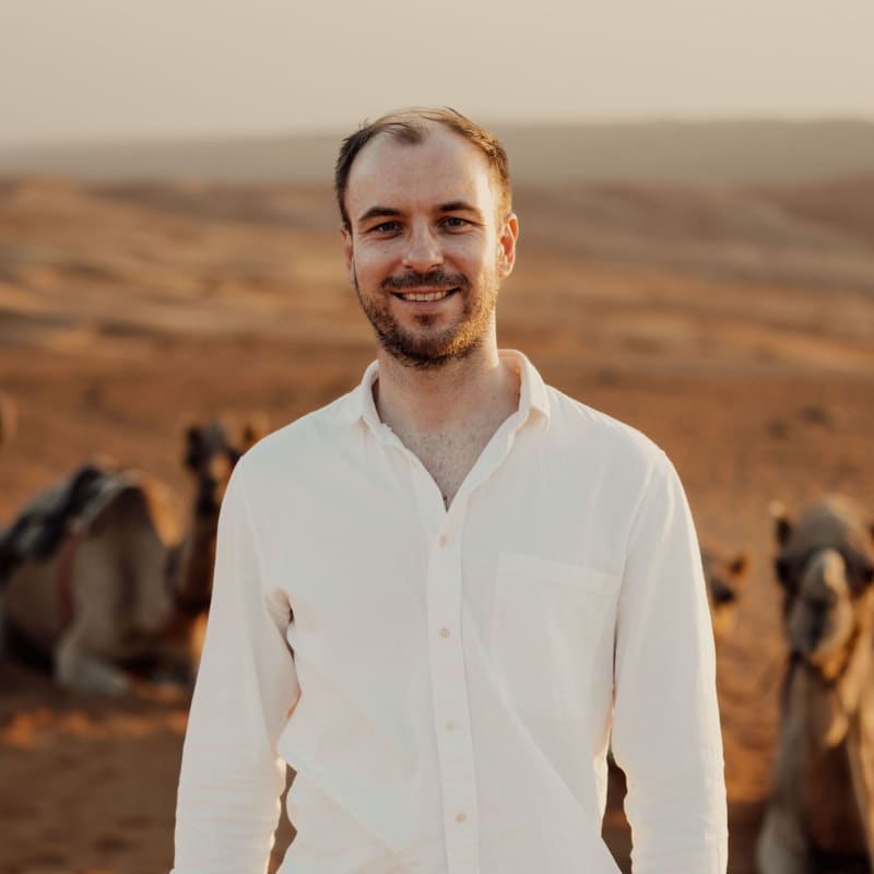 A smiling man in a white shirt standing in a desert with camels in the background