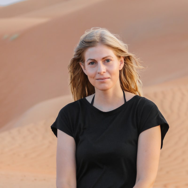 A woman with blonde hair stands in a desert landscape with sand dunes behind her.