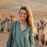 A young woman with long, wavy hair smiling outdoors in a desert-like setting with camels in the background.
