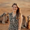 A young woman with long brown hair smiling outdoors with camels in the background.