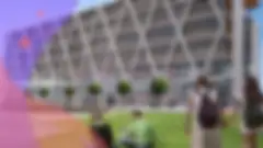 a male and female college student sit on a green lawn outside a futuristic building with a row of trees in front of it while another student wearing a backpack is walking nearby