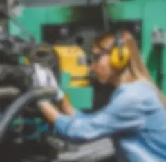 Frau mit Gehörschutz, Augenschutz und Handschuhen bei der Arbeit an einer Industriemaschine.