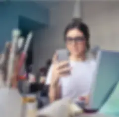 Eine junge Frau mit Brille und weißem T-Shirt schaut auf ein Handy, während sie an einem Kunsttisch mit Bechern voller Buntstifte und Pinsel steht. Vor ihr steht ein geöffneter Laptop.