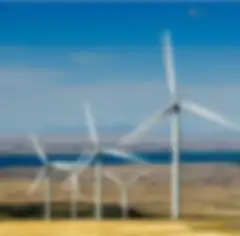 A row of wind turbines on a hill with yellow grass, a blue sky with wispy clouds, a reservoir in the background, and hazy mountains in the distance