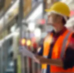 A man wearing a hard hat and safety vest is holding a clipboard and looking up at inventory on shelves in a warehouse.