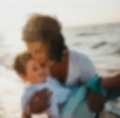 A woman with sunglasses on her head holding a young boy laughing on the shore of a beach