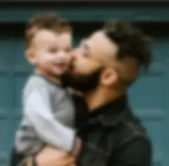 Man with curly hair and glasses holding a toddler boy with curly hair in his arms and kissing him on the cheek