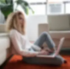 Woman with curly hair and reading glasses reclining against sofa working on a laptop