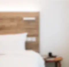 A cropped view of a hotel bed with white linens and a natural wood plank headboard. There is a small, round, natural wood table beside the bed, against a white wall.