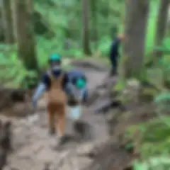 Three people whose faces are not visible, wearing hard hats and work clothes, carry a bucket on a dirt trail in a forest