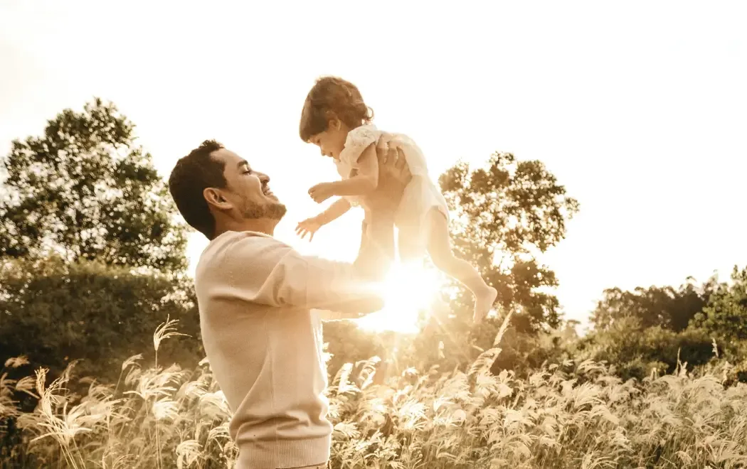 A man wearing a light sweater lifts a smiling child in the air in a field with tall grass and trees at sunset.