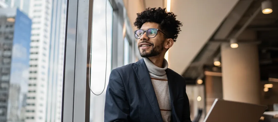 A person with glasses and curly hair, wearing a turtleneck and blazer, sits by a window with a laptop, looking outside thoughtfully in a modern office.