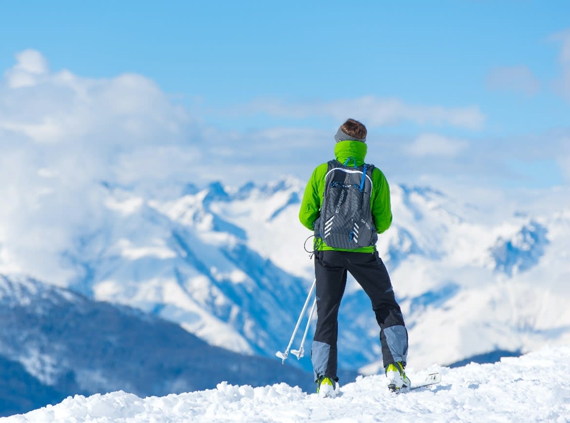 Photo of a person, facing away from the camera, wearing a bright green ski jacket and black ski pants standing on snow covered ground viewing snow covered mountain tops all around them