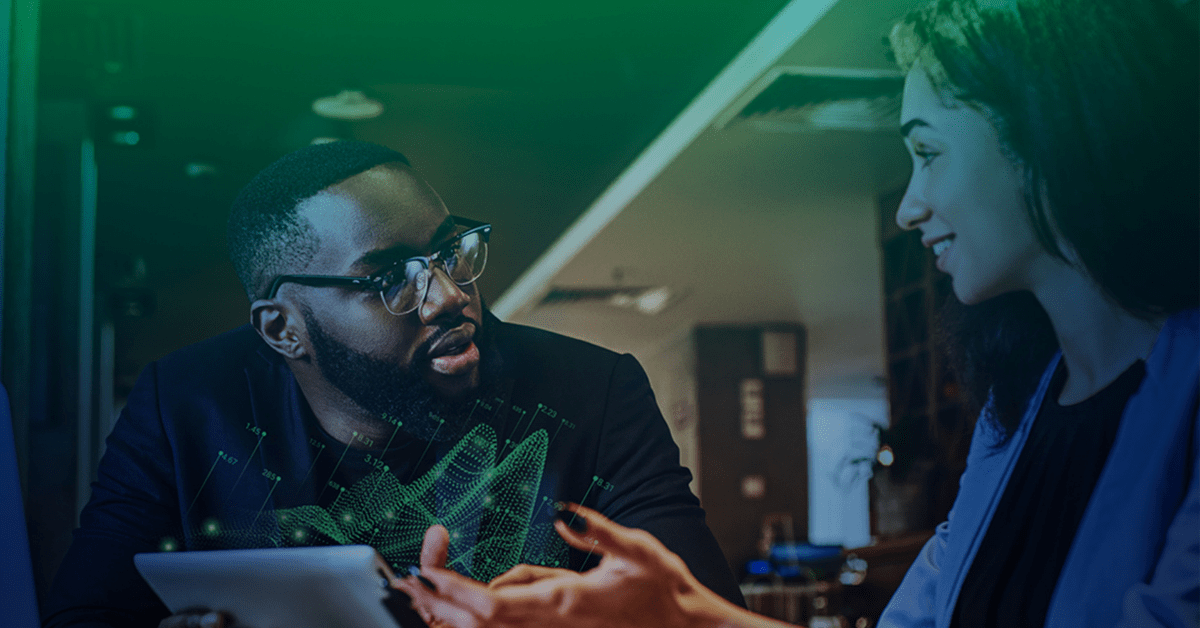 Photo of a man and a woman in an office space having a conversation with a laptop open on the table in front of them