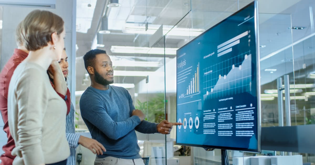 Photo of four people in a conference room, gathered around a television screen displaying charts and graphs, discussing what they are viewing