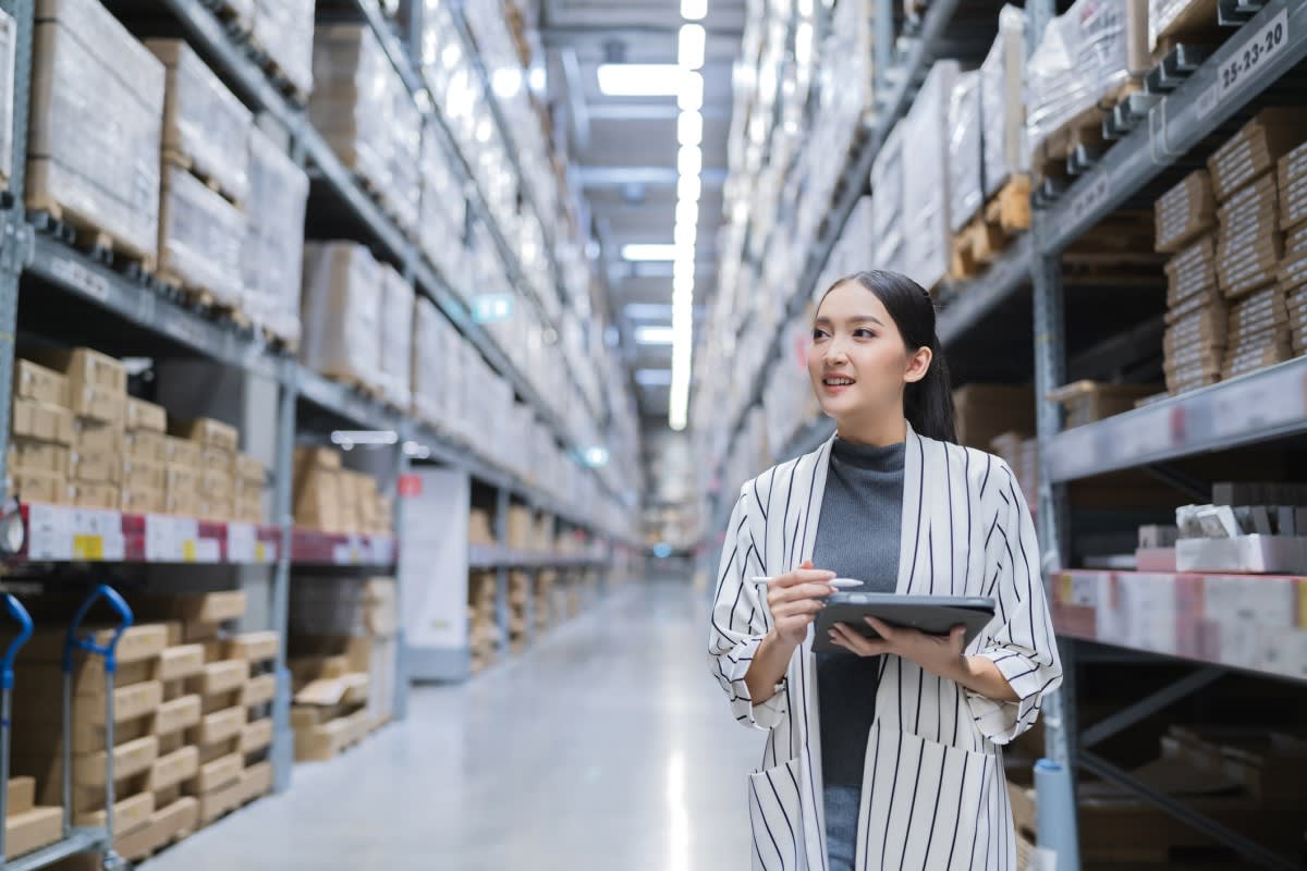 Photo of a woman holding a tablet reviewing warehouse shelves with boxes and products on them