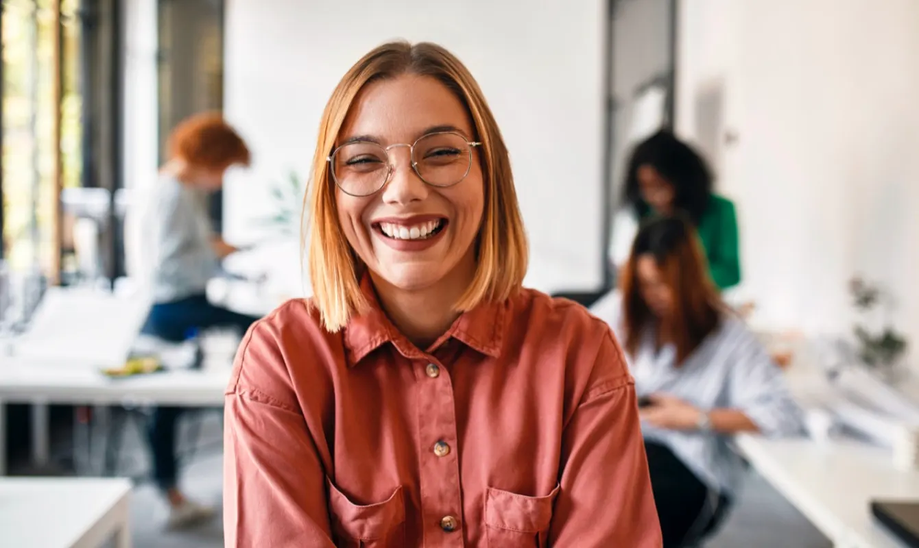 A smiling woman with glasses and shoulder-length hair, wearing a red shirt, stands in focus in an office setting with three people blurred in the background.