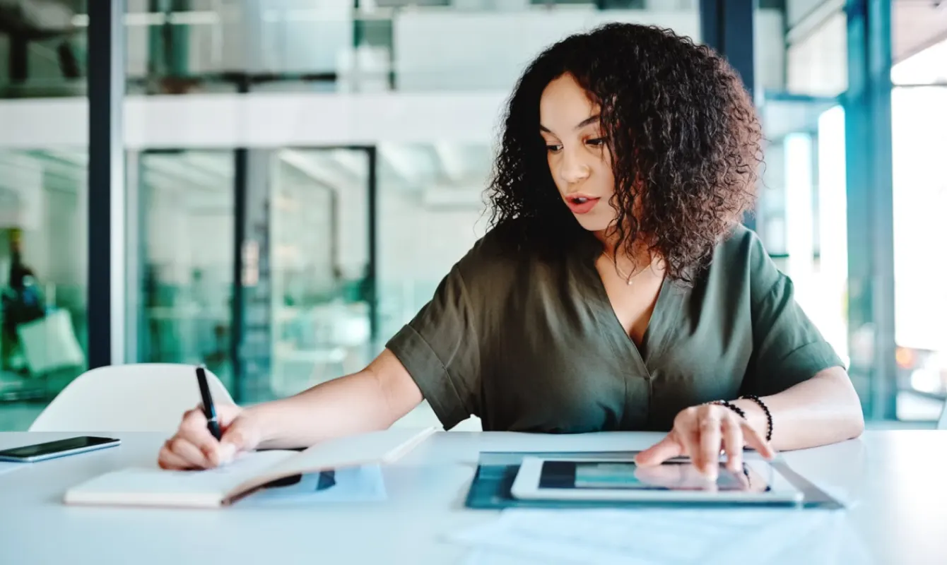 A person with curly hair, wearing a green shirt, sits at a desk. They are writing in a notebook with one hand and using a tablet with the other. A smartphone is also on the desk.