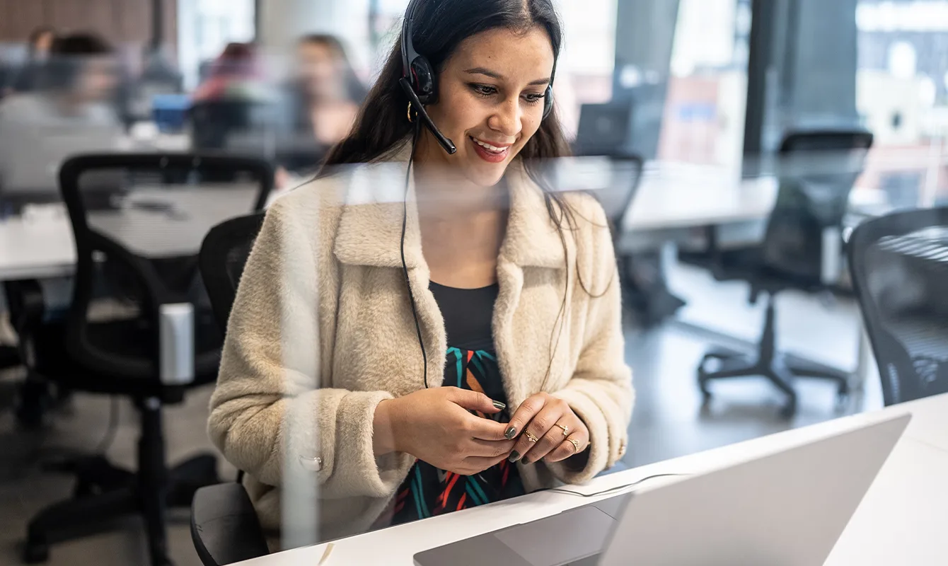 Mulher usando headset e um casaco de cor clara sentada em uma mesa com notebook e trabalhando em um escritório com outras pessoas desfocadas ao fundo.