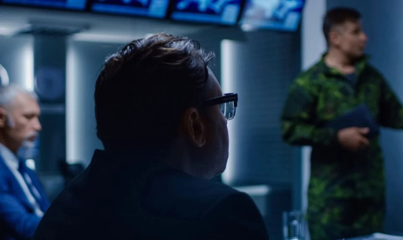 A man in glasses interacts with others in a dimly lit room. A camouflaged military officer stands and presents information, while another seated man observes. Multiple screens are visible in the background.