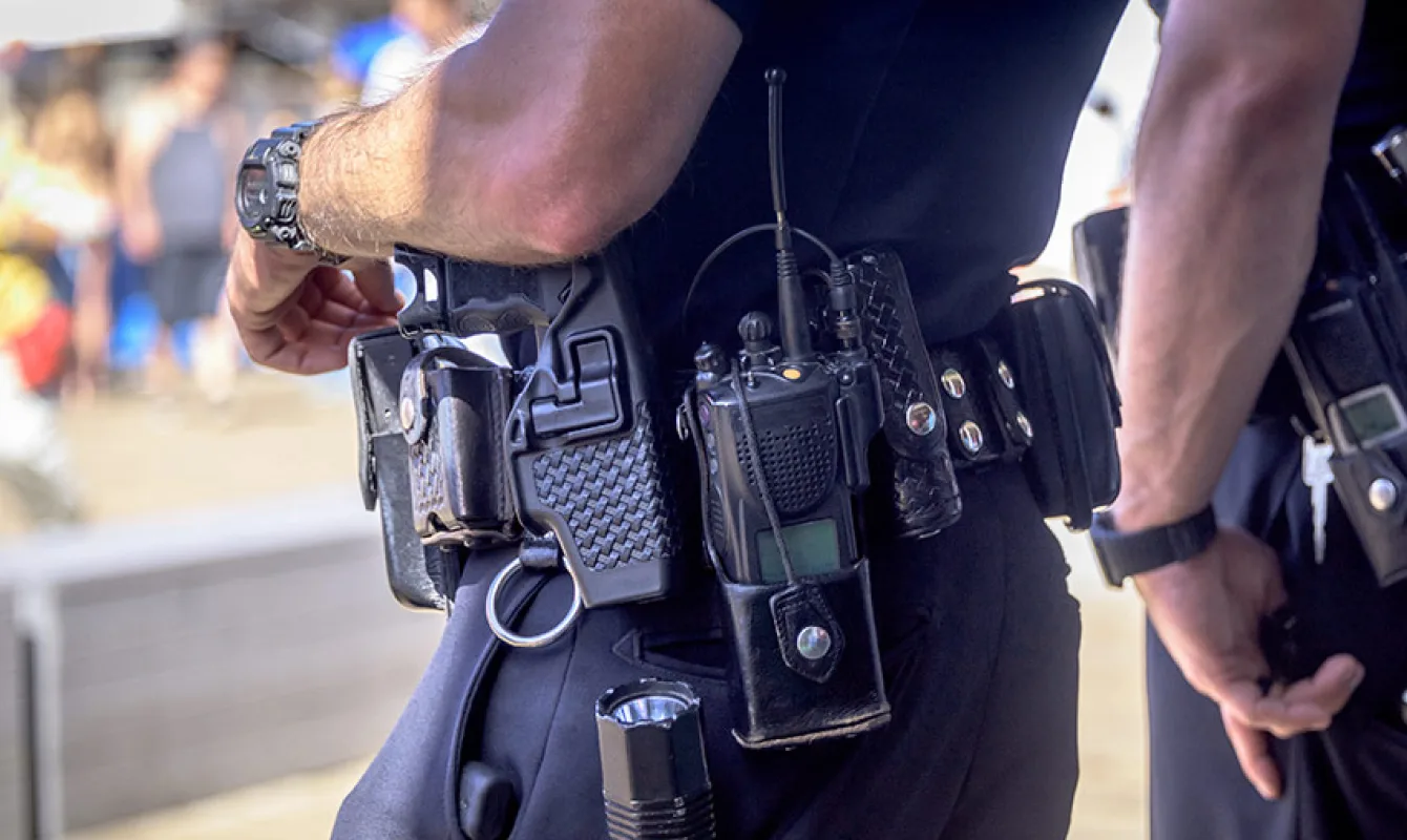 Close-up of two police officers' belts equipped with hand radios, handcuffs, and holstered firearms, with blurred background.