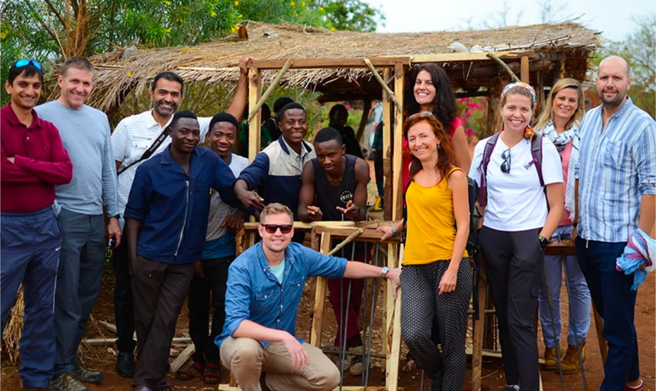 A diverse group of people is posing and smiling in front of a small thatched structure in an outdoor, rural setting.