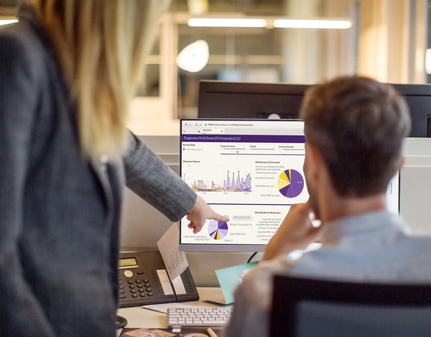 Two people at a desk look at a computer screen displaying charts and graphs. One person points to a pie chart while holding a printed version of the same chart.