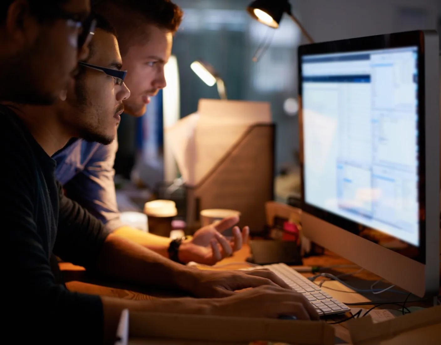 Three people are working late at night, intently looking at a computer screen displaying a document editor. They are surrounded by papers and illuminated by desk lamps.