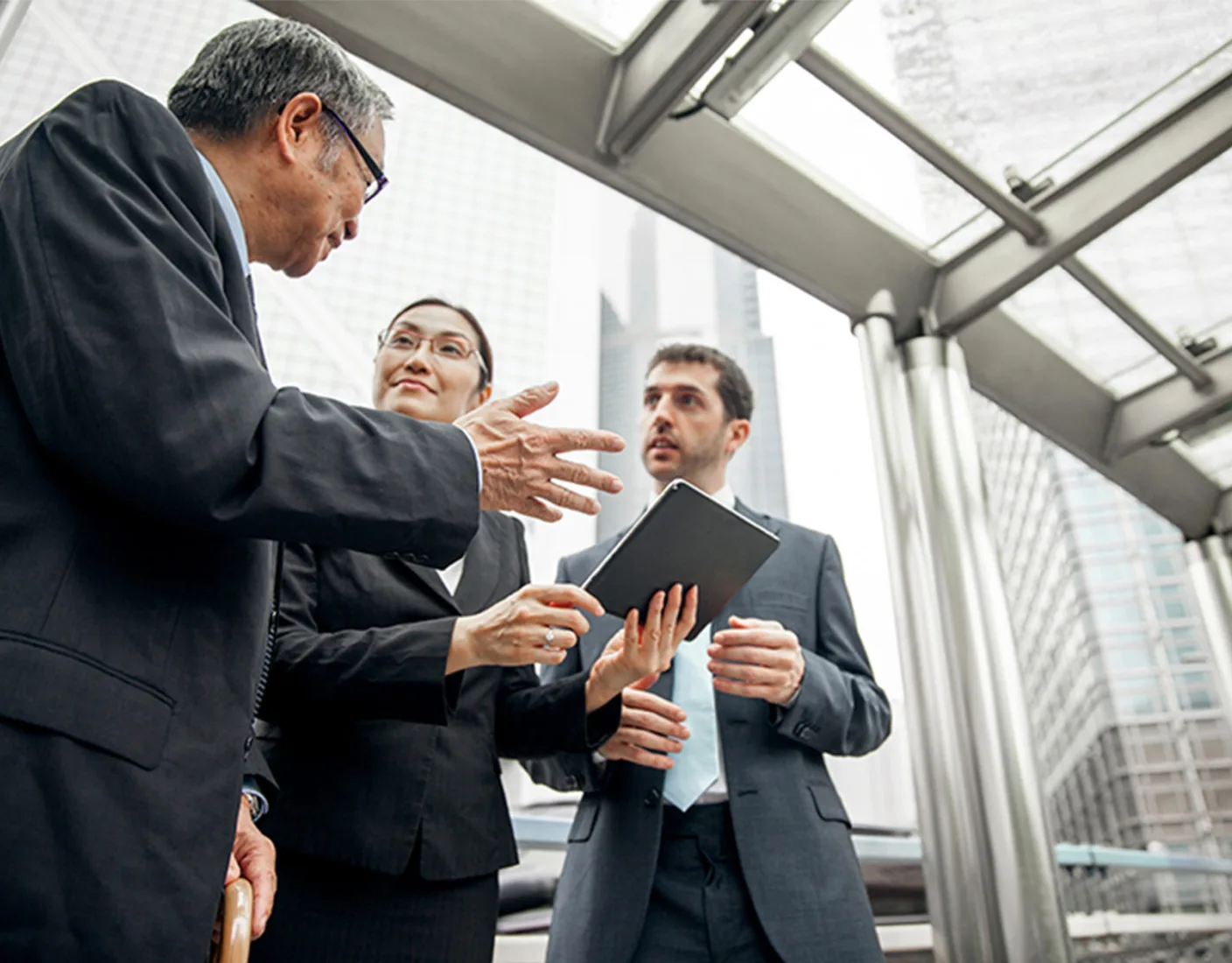 Three business professionals in suits engage in a discussion, with one holding a tablet. The conversation takes place in an outdoor urban setting with tall buildings visible in the background.