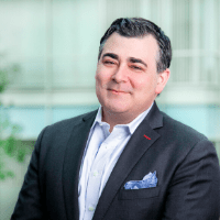 Headshot of blog author Joe DosSantos. He has dark hair, is wearing a dark suit, white shirt, and a pocket square stands smiling in front of a blurred background.