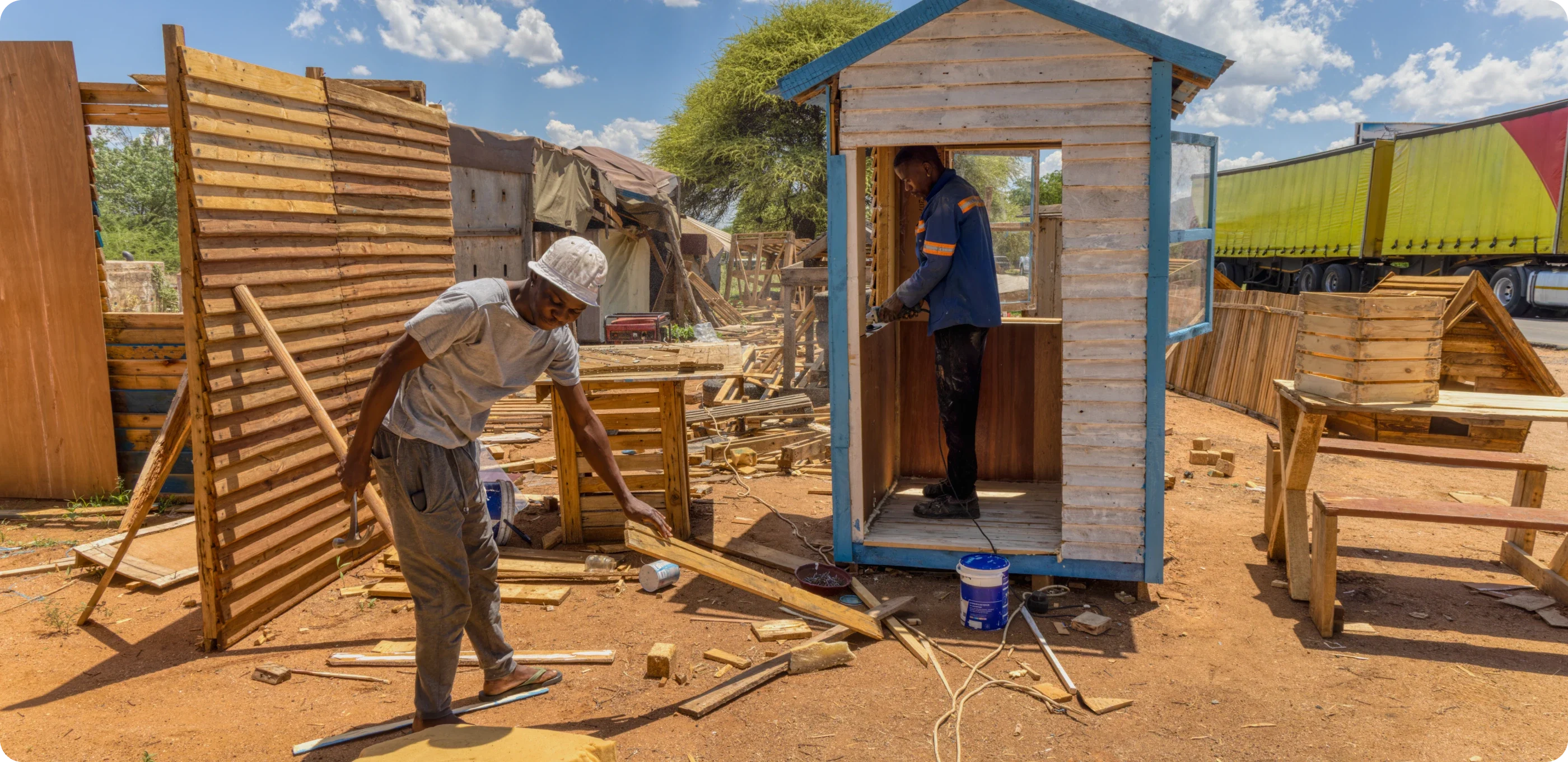 Two men work outdoors on a construction site, assembling wooden structures under a sunny sky. One man is inside a small wooden building, while the other measures and cuts wood outside.