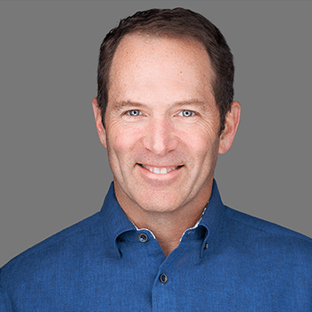 Headshot of blog author, Drew Clarke. He has short brown hair, wearing a blue button-up shirt, and is smiling in front of a plain gray background.