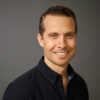 Headshot of blog author Ryan Welsh. He has short, light brown hair and blue eyes, is smiling, wearing a dark button-up shirt against a plain gray background.