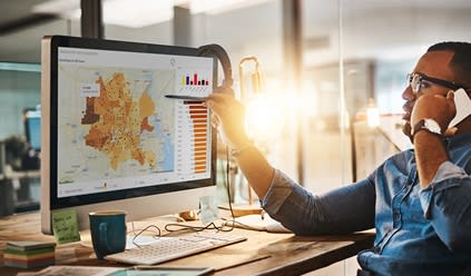 A man in glasses speaks on the phone while analyzing data on a computer screen displaying a map and charts in an office.
