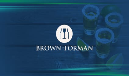 Brown-Forman logo with a white background. Behind the logo are four filled shot glasses with rims dipped in salt, positioned next to lime slices on a dark wooden table.