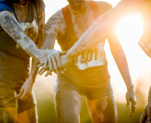 Three people covered in mud stack their hands together in a gesture of teamwork. The sun is shining brightly in the background.