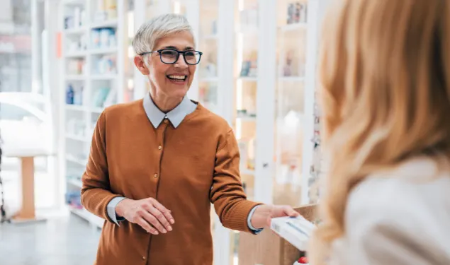 A person with short gray hair and glasses hands an item to another person in a store setting. Shelves with various products are visible in the background.